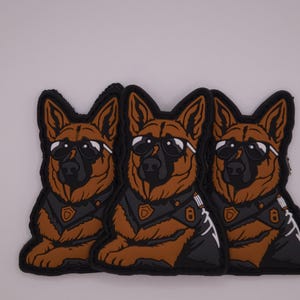 May include: Three patches featuring a German Shepherd dog wearing sunglasses and a tactical vest. The patches are brown, black, and white, with a detailed design. The dog is in a seated position.