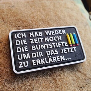 Könnte beinhalten: Ein rechteckiger schwarzer Aufnäher mit weißem Text: "ICH HAB WEDER DIE ZEIT NOCH DIE BUNTSTIFTE UM DIR DAS JETZT ZU ERKLÄREN...". Er hat einen weißen Rand und drei kleine Buntstift-Grafiken.