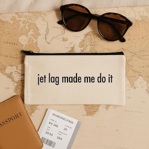 May include: A cream-coloured travel pouch with the text "jet lag made me do it" in black. Tortoiseshell sunglasses rest above the pouch on a world map. A passport and boarding pass are also visible.