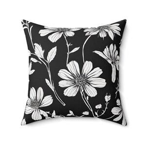 May include: Square throw pillow with a black background and a white floral pattern. The design features large, stylized flowers and leaves, creating a striking contrast. The pillow is a decorative home accent.