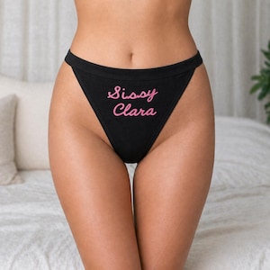 Peut inclure: Culotte noire avec l'inscription rose "Sissy Clara". Le sous-vêtement est de coupe haute et de conception simple. En arrière-plan, un lit avec des oreillers blancs et une plante.