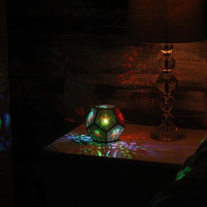 Pu&ograve; includere: Una lampada geometrica multicolore proietta motivi colorati su una superficie e una parete. Una lampada in vetro trasparente con paralume in tessuto &egrave; accanto alla lampada, creando un'atmosfera calda.