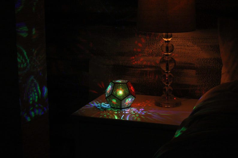 Pu&ograve; includere: Una lampada geometrica multicolore proietta motivi colorati su una superficie e una parete. Una lampada in vetro trasparente con paralume in tessuto &egrave; accanto alla lampada, creando un'atmosfera calda.