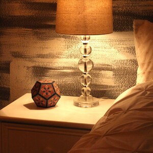 Pu&ograve; includere: Una scena da comodino con una lampada in vetro trasparente con paralume beige e un oggetto decorativo geometrico multicolore. La lampada &egrave; accesa, proiettando un bagliore caldo sul comodino bianco e sull'area circostante. Un letto con una trapunta &egrave; parzialmente visibile.