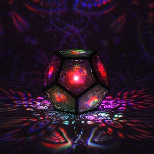 Pu&ograve; includere: Una lampada geometrica sfaccettata con intricati motivi floreali. La lampada proietta motivi di luce colorati sulla superficie circostante. Il design della lampada presenta una sorgente luminosa rossa centrale, con sfumature verdi, rosse e blu.