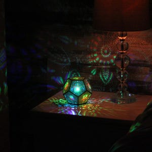 Pu&ograve; includere: Un apparecchio di illuminazione geometrico multicolore &egrave; posto su un comodino, proiettando motivi colorati sulle superfici circostanti. &Egrave; visibile anche una lampada con base in vetro e paralume. La scena &egrave; debolmente illuminata, creando un'atmosfera calda.