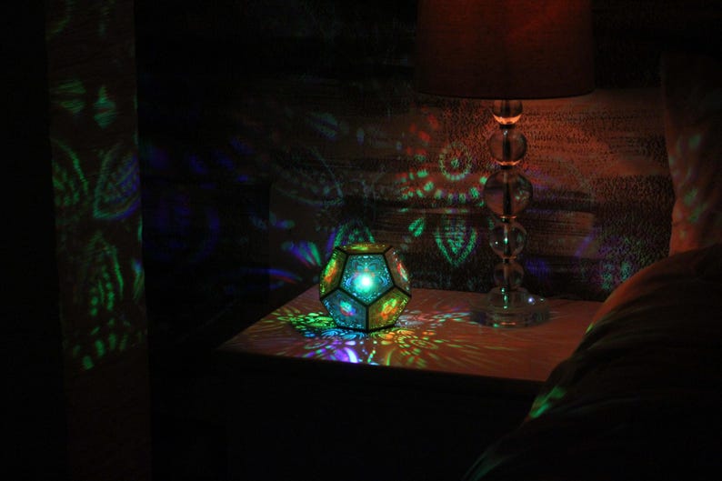 Pu&ograve; includere: Un apparecchio di illuminazione geometrico multicolore &egrave; posto su un comodino, proiettando motivi colorati sulle superfici circostanti. &Egrave; visibile anche una lampada con base in vetro e paralume. La scena &egrave; debolmente illuminata, creando un'atmosfera calda.