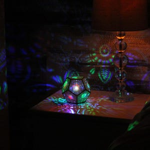 Pu&ograve; includere: Un apparecchio di illuminazione geometrico multicolore che proietta motivi colorati su una superficie. Sullo sfondo c'&egrave; una lampada con un paralume marrone. La stanza &egrave; buia, con l'apparecchio di illuminazione come fonte di luce primaria.