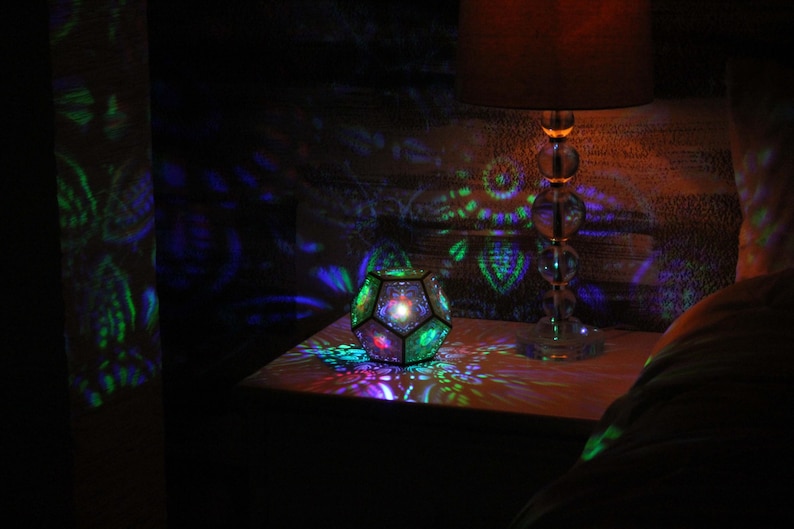 Pu&ograve; includere: Un apparecchio di illuminazione geometrico multicolore che proietta motivi colorati su una superficie. Sullo sfondo c'&egrave; una lampada con un paralume marrone. La stanza &egrave; buia, con l'apparecchio di illuminazione come fonte di luce primaria.