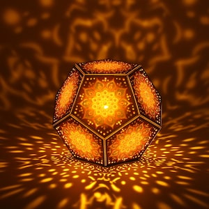 May include: A dodecahedron-shaped lamp featuring intricate laser-cut mandala patterns on each pentagonal face. The lamp emits a warm, golden light, casting detailed, symmetrical floral-like shadows onto the surrounding area from its internal source.