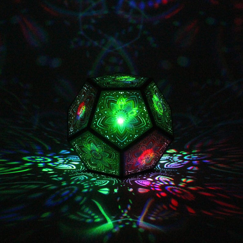 Pu&ograve; includere: Lampada geometrica sfaccettata con intricati motivi floreali. La lampada emette una vivace luce verde dall'interno, proiettando ombre colorate sulla superficie. Il design presenta una miscela di tonalit&agrave; verdi, rosse e blu, creando un effetto psichedelico.