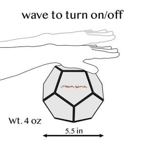 Pu&ograve; includere: Un oggetto geometrico grigio chiaro con finiture nere, con il testo "MKHA WMA" e "wave to turn on/off". Misura 14 cm di larghezza e pesa 113 grammi.