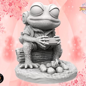 Pode incluir: Uma figura impressa em 3D de cor cinza de um sapo usando uma mochila e segurando um pequeno objeto em suas mãos. O sapo está sentado em uma rocha com uma fogueira atrás.