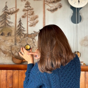 May include: A woman in a blue sweater holds a small, round jar with a yellow lid. The jar is in focus, and the woman's face is out of focus. The background is a wall with a painted forest scene.