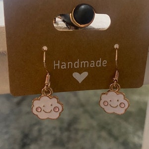 May include: A pair of gold-tone earrings with a white cloud charm featuring a pink smiley face. The earrings are displayed on a brown card with the word "Handmade" written on it.