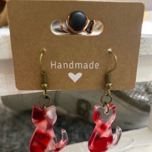 Puede incluir: Pendientes de gato hechos a mano de color rojo y blanco con un acabado de resina transparente. Los pendientes están en una tarjeta marrón con la palabra "Handmade" y un corazón.