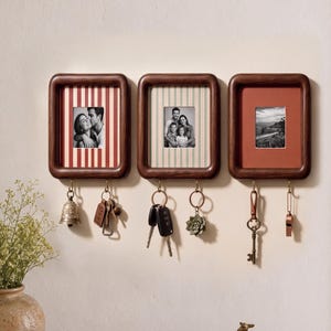 May include: Three rectangular dark wood wall-mounted frames with rounded corners, each featuring a central photo and two metal hooks underneath. The frames display red striped, green striped, and solid terracotta backings with various hanging keys.
