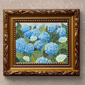 May include: A framed painting of blue hydrangeas in a gold frame. The painting depicts a cluster of blue hydrangeas with green leaves and white flowers.