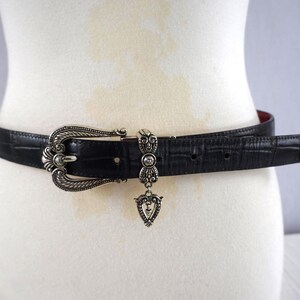 K&ouml;nnte beinhalten: Schwarzer G&uuml;rtel mit silberner Schnalle und dekorativen Elementen. Der G&uuml;rtel hat eine strukturierte Oberfl&auml;che und ein rotes Innenfutter. Ein silberner Anh&auml;nger h&auml;ngt in der Mitte.