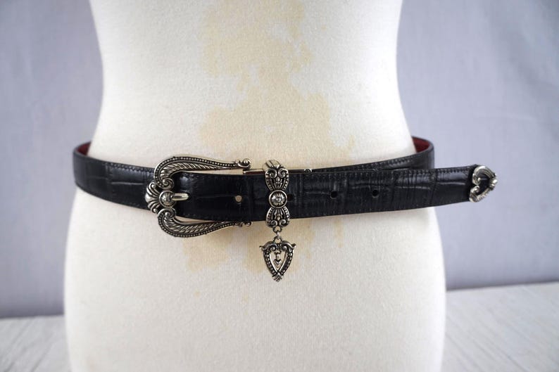 K&ouml;nnte beinhalten: Schwarzer G&uuml;rtel mit silberner Schnalle und dekorativen Elementen. Der G&uuml;rtel hat eine strukturierte Oberfl&auml;che und ein rotes Innenfutter. Ein silberner Anh&auml;nger h&auml;ngt in der Mitte.