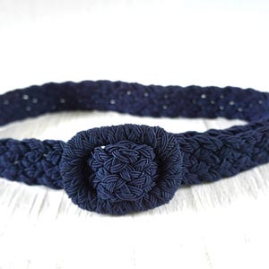 May include: A navy blue braided belt with a round, knotted buckle. The belt is made of a thick, woven material and is displayed on a white surface. The belt is a fashion accessory.