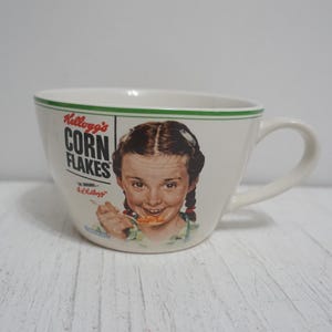 May include: White ceramic mug with a green rim and a picture of a girl eating Kellogg's Corn Flakes. The text on the mug reads "Kellogg's Corn Flakes The Original K. Kellogg's".