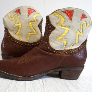 K&ouml;nnte beinhalten: Braune Leder-Cowgirl-Stiefel mit einem wei&szlig;en und goldenen geometrischen Muster am oberen Teil des Stiefels.