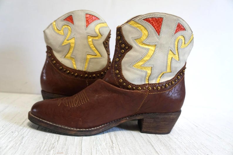 K&ouml;nnte beinhalten: Braune Leder-Cowgirl-Stiefel mit einem wei&szlig;en und goldenen geometrischen Muster am oberen Teil des Stiefels.
