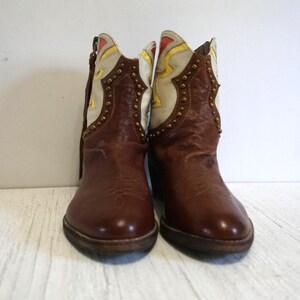 K&ouml;nnte beinhalten: Ein Paar braune Leder-Cowbo Stiefel mit wei&szlig;en und gelben Akzenten. Die Stiefel haben dekorative Nieten und eine spitze Zehenpartie.