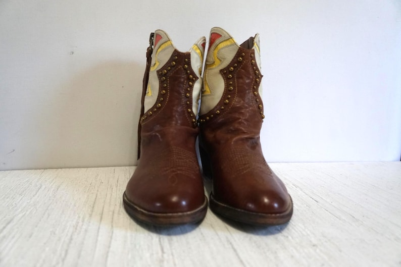 K&ouml;nnte beinhalten: Ein Paar braune Leder-Cowbo Stiefel mit wei&szlig;en und gelben Akzenten. Die Stiefel haben dekorative Nieten und eine spitze Zehenpartie.