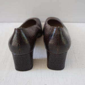 May include: A pair of brown leather heeled shoes with a textured finish. The shoes have a low block heel and a closed toe.
