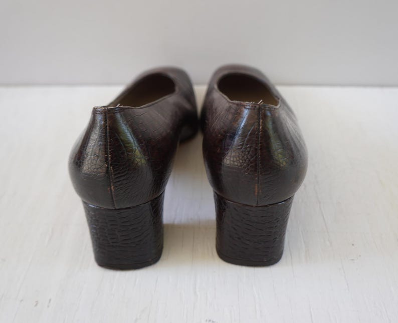 May include: A pair of brown leather heeled shoes with a textured finish. The shoes have a low block heel and a closed toe.