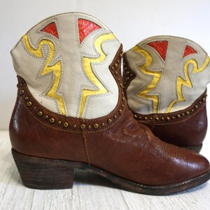 K&ouml;nnte beinhalten: Braune Leder-Cowgirl-Stiefel mit einem wei&szlig;en und gelben geometrischen Muster auf der Oberseite. Die Stiefel haben eine spitze Zehenpartie und einen kleinen Absatz.