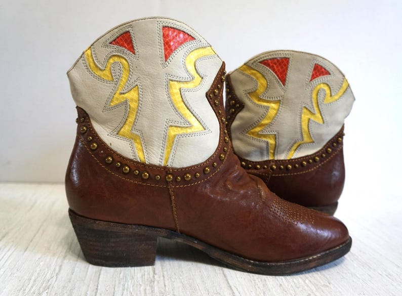 K&ouml;nnte beinhalten: Braune Leder-Cowgirl-Stiefel mit einem wei&szlig;en und gelben geometrischen Muster auf der Oberseite. Die Stiefel haben eine spitze Zehenpartie und einen kleinen Absatz.