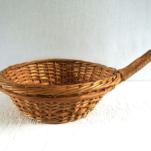 May include: A round, light brown woven wicker basket with a single long, sturdy handle extending from one side. The basket features a tight, intricate weave pattern throughout the bowl and the handle, creating a functional, open-top container.