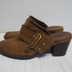 May include: Brown leather heeled clogs with a buckle detail. The clogs have a wooden heel and a leather strap with a metal buckle.