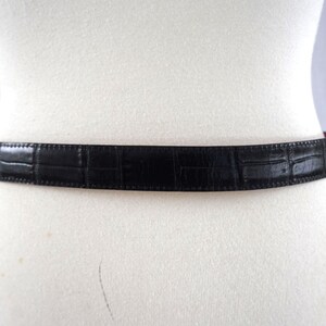 K&ouml;nnte beinhalten: Ein schwarzer G&uuml;rtel mit Krokodilhautmuster wird auf einer wei&szlig;en Schaufensterpuppe pr&auml;sentiert. Der G&uuml;rtel besteht aus einem strukturierten Material und hat ein klassisches Design. Der G&uuml;rtel ist ein Modeaccessoire.