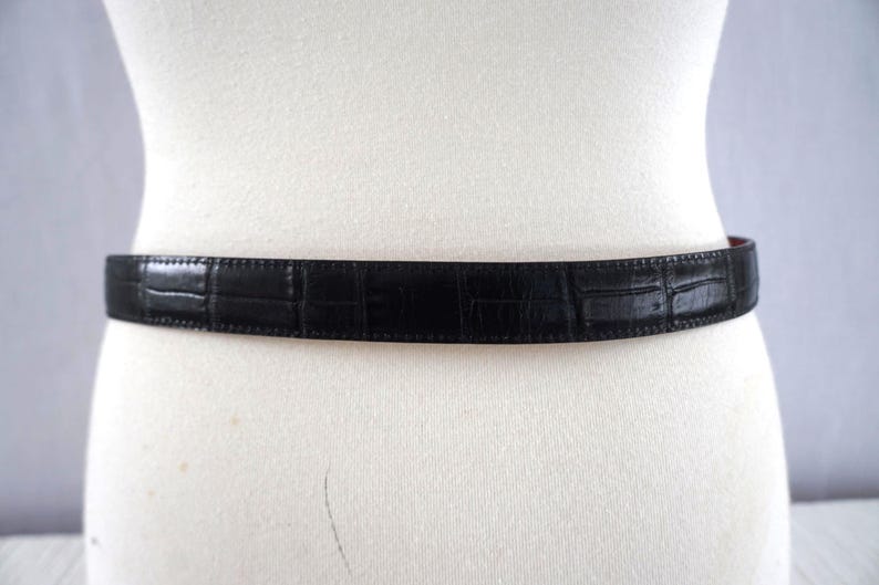 K&ouml;nnte beinhalten: Ein schwarzer G&uuml;rtel mit Krokodilhautmuster wird auf einer wei&szlig;en Schaufensterpuppe pr&auml;sentiert. Der G&uuml;rtel besteht aus einem strukturierten Material und hat ein klassisches Design. Der G&uuml;rtel ist ein Modeaccessoire.