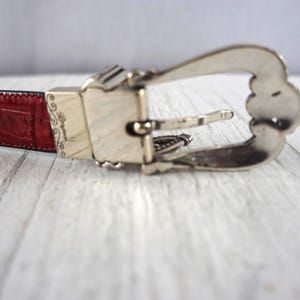 K&ouml;nnte beinhalten: Nahaufnahme eines roten Lederg&uuml;rtels mit silberner Schnalle. Der G&uuml;rtel hat ein strukturiertes, krokodilartiges Muster. Die Schnalle ist mit dekorativen Details verziert.