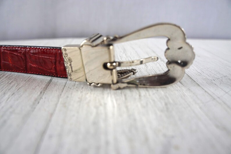 K&ouml;nnte beinhalten: Nahaufnahme eines roten Lederg&uuml;rtels mit silberner Schnalle. Der G&uuml;rtel hat ein strukturiertes, krokodilartiges Muster. Die Schnalle ist mit dekorativen Details verziert.