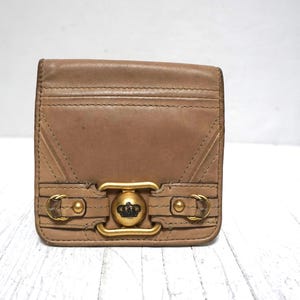 May include: A tan leather wallet with a gold-tone clasp and a crown emblem. The wallet has a rectangular shape with stitched details and a flap closure. The hardware includes a decorative buckle and rivets.