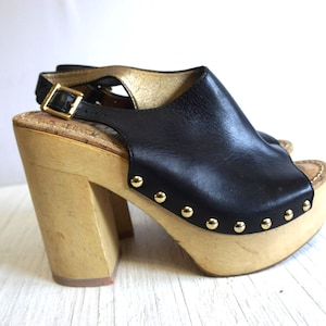 May include: Black leather platform sandals with a wooden base and heel. The sandals have a slingback strap with a gold buckle and are adorned with gold studs along the platform. The heel height is approximately 4 inches.