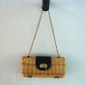May include: A rectangular woven wicker handbag with a black flap and gold-tone clasp. The bag has a chain strap and small metal feet. The bag is hanging against a white background.