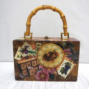 May include: A vintage-style box purse with a bamboo handle and gold-tone hardware. The box is decorated with floral and botanical imagery, including a clock face, stamps, and flowers. The color palette includes brown, beige, and pink.