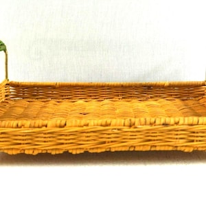 May include: A rectangular yellow woven wicker tray featuring two gold-toned metal handles with green woven accents. The tray is supported by four small gold-toned metal feet, and the handles have a decorative curled design at each end.
