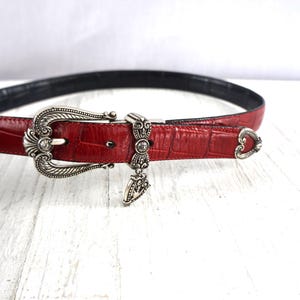 K&ouml;nnte beinhalten: Ein roter Lederg&uuml;rtel mit Krokodiltextur. Der G&uuml;rtel hat eine silberfarbene Schnalle mit aufwendigen Details und einem Anh&auml;nger. Die Innenseite des G&uuml;rtels ist schwarz, und die Schnalle hat einen kleinen, klaren Edelstein.