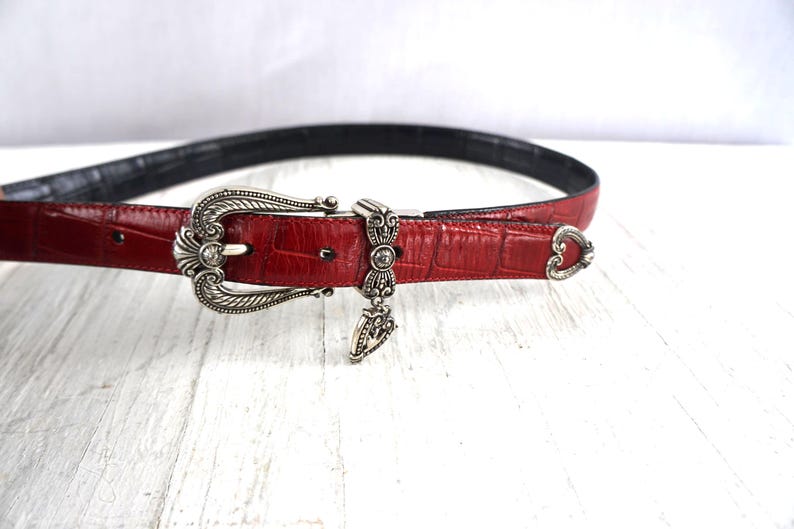 K&ouml;nnte beinhalten: Ein roter Lederg&uuml;rtel mit Krokodiltextur. Der G&uuml;rtel hat eine silberfarbene Schnalle mit aufwendigen Details und einem Anh&auml;nger. Die Innenseite des G&uuml;rtels ist schwarz, und die Schnalle hat einen kleinen, klaren Edelstein.