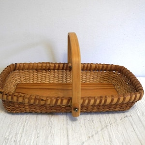 May include: Rectangular woven basket with a wooden base and a tall, arched handle. The basket is made of light brown wicker and has a sturdy, handcrafted appearance. The handle is attached with a metal rivet. Ideal for storage or display.