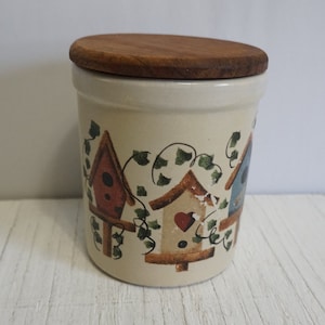 May include: A ceramic jar with a wooden lid, featuring a hand-painted design of three birdhouses with ivy vines. The birdhouses are painted in shades of red, blue, and brown.
