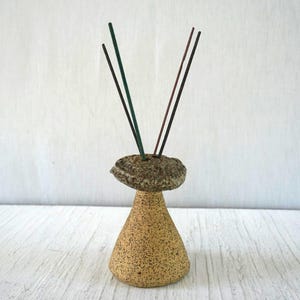 May include: A ceramic incense holder with a speckled tan conical base and a textured, stone-like top piece. Four thin incense sticks in shades of green and brown are inserted into the top, extending upwards in a fan-like arrangement.